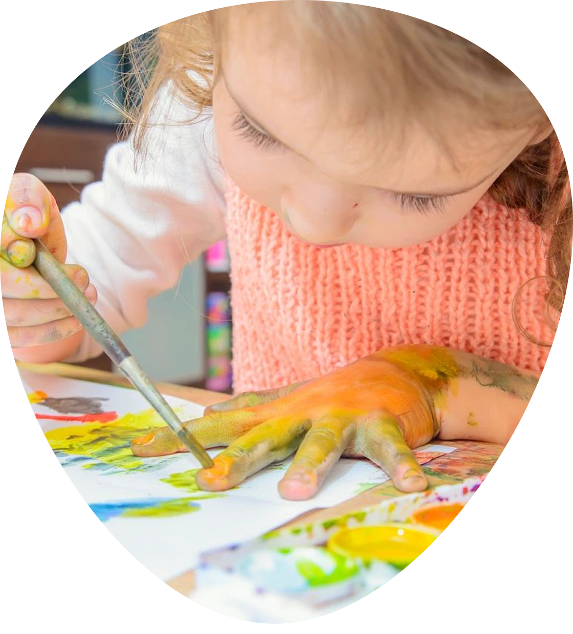 Group 1321317167 Child painting with a brush, hand covered in multiple colors. Wearing a white shirt and pink sweater, focused on artwork on the table.