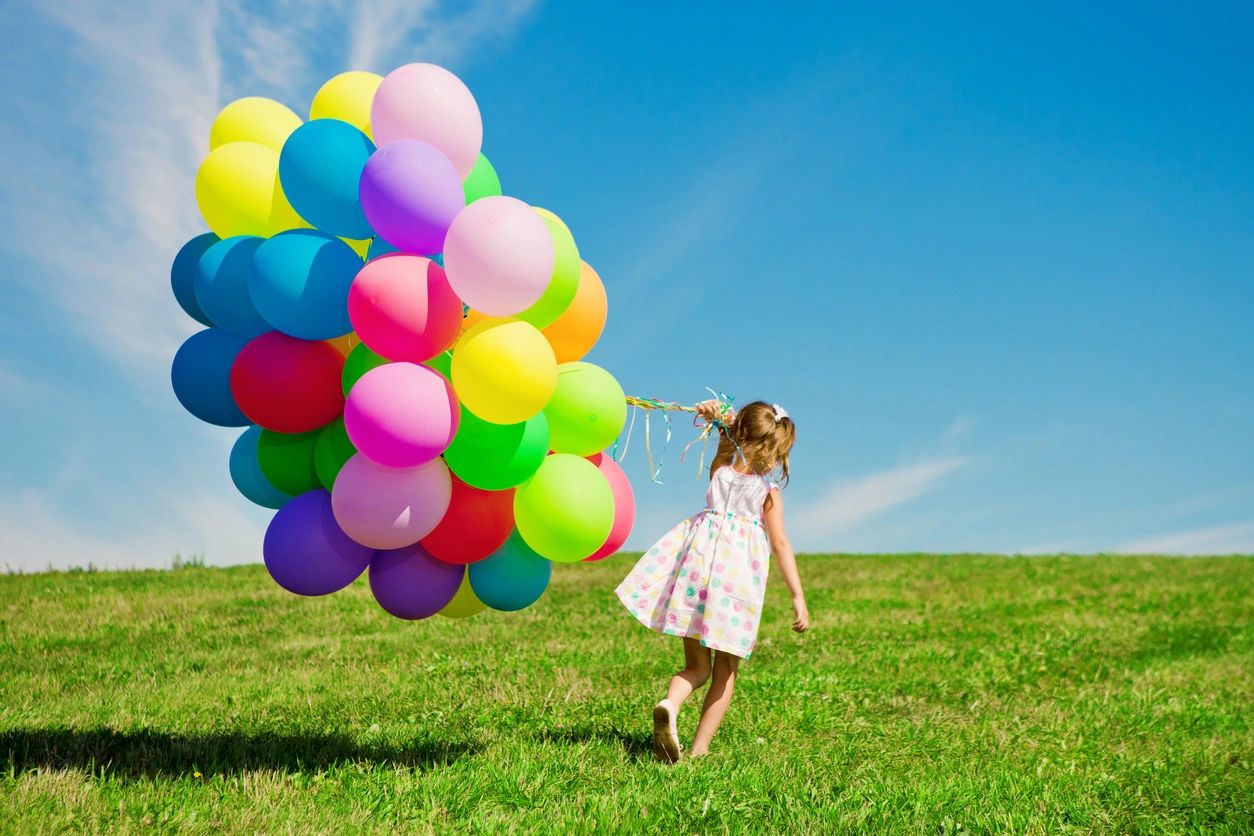 fm=f_JGrdwKj A child in a polka dot dress holds a large bunch of colorful balloons while walking on a grassy hill under a clear blue sky.