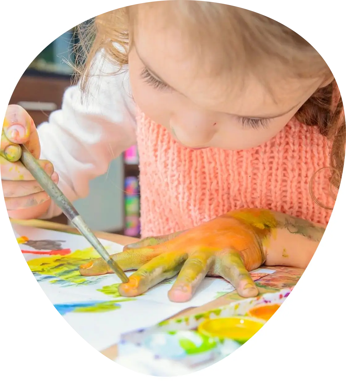 Child painting with a brush, hand covered in multiple colors. Wearing a white shirt and pink sweater, focused on artwork on the table.